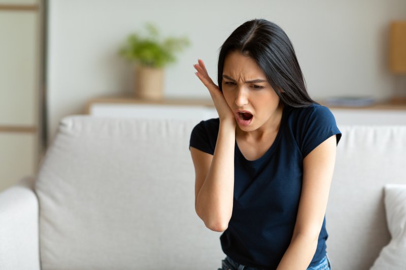 A woman seated on a couch and holding her jaw while cringing in pain