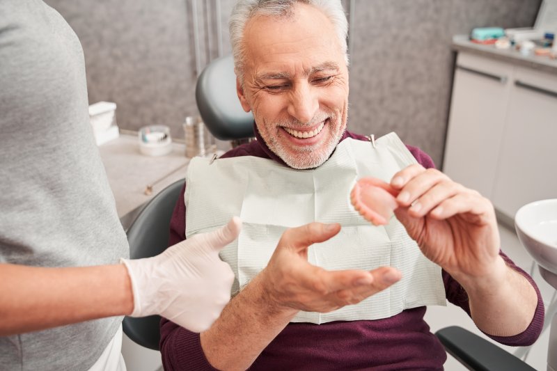Older man in dental chair smiling at dentures in his hand as dentist off to the side gives gloved thumbs up